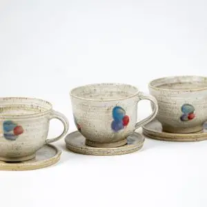 כוסות קרמיקה בעבודת יד על צלוחיות תואמות, עם דוגמאות מופשטות של כתמי כחול ואדום.