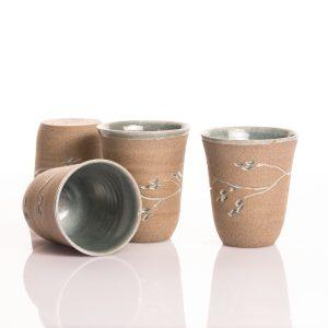 כוס קרמיקה גבוהה וצרה בצבע בז' עם מרקם חולי, עיטור ענף מגולף ופנים בגלזורה אפורה-תכולה.