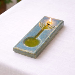 פמוט זיכרון מקרמיקה בעבודת יד, בגווני ירוק-כחול עם עיצוב בהשראת מנורה, מונח על מפה לבנה. במרכזו פתיל דולק המפיץ אור רך וחמים, תוך הדגשת הברק והמרקם של הגלזורה.